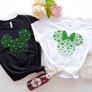 May include: Two t-shirts with a green shamrock design in the shape of Mickey Mouse ears. One shirt is black and the other is white. The white shirt has a green bow on the ears.