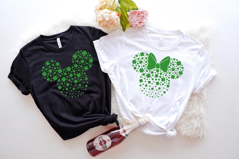 May include: Two t-shirts with a green shamrock design in the shape of Mickey Mouse ears. One shirt is black and the other is white. The white shirt has a green bow on the ears.