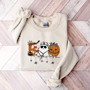May include: Cream-colored sweatshirt featuring a Halloween-themed graphic. The design includes a spider, a bat, a ghost wearing a nurse's cap, and a jack-o'-lantern. The graphic is in shades of orange, black, and white, perfect for the spooky season.