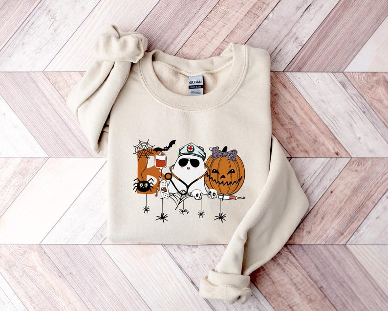 May include: Cream-colored sweatshirt featuring a Halloween-themed graphic. The design includes a spider, a bat, a ghost wearing a nurse's cap, and a jack-o'-lantern. The graphic is in shades of orange, black, and white, perfect for the spooky season.