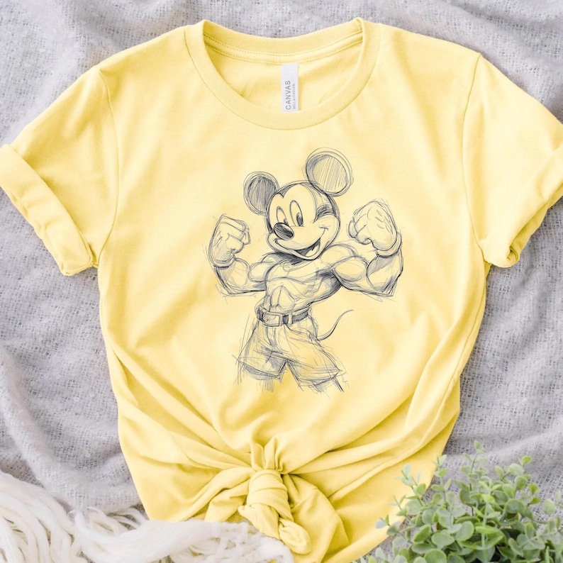 May include: A light yellow short-sleeved t-shirt featuring a dark grey sketch-style graphic of a muscular cartoon mouse character with large round ears, flexing both arms, wearing shorts and a belt, with the word CANVAS printed on the inner neck label.