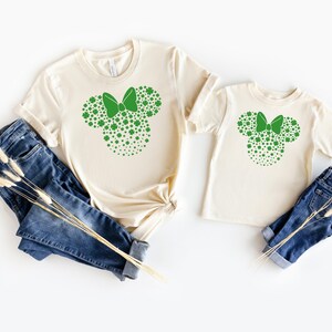 May include: Two cream-colored t-shirts with a green shamrock design in the shape of a bow. The design is a silhouette of a mouse head with a bow.