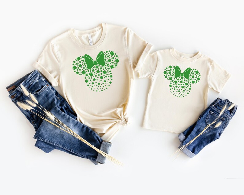May include: Two cream-colored t-shirts with a green shamrock design in the shape of a bow. The design is a silhouette of a mouse head with a bow.