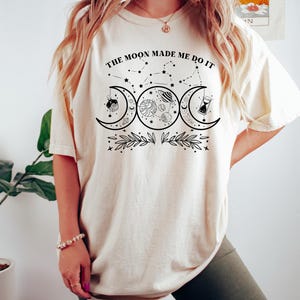 May include: A white t-shirt with a black graphic design featuring three moons, stars, planets, and the text "The Moon Made Me Do It".