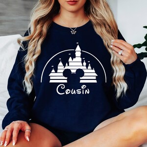 May include: A navy blue sweatshirt featuring a white graphic of a castle and Mickey Mouse ears. The text "Cousin" is printed below the graphic.