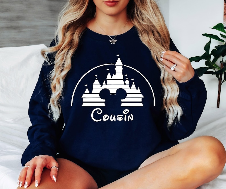 May include: A navy blue sweatshirt featuring a white graphic of a castle and Mickey Mouse ears. The text "Cousin" is printed below the graphic.