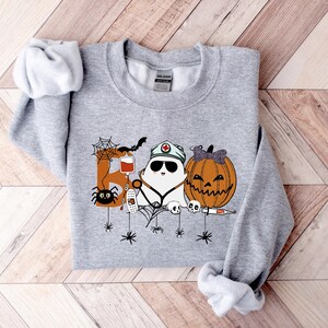May include: A light gray sweatshirt with a Halloween-themed graphic. The graphic features a ghost wearing a nurse's hat, a carved pumpkin, and the word "Boo" with spiders and other Halloween elements.