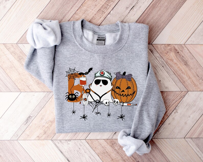 May include: A light gray sweatshirt with a Halloween-themed graphic. The graphic features a ghost wearing a nurse's hat, a carved pumpkin, and the word "Boo" with spiders and other Halloween elements.