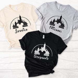 May include: Three t-shirts featuring a black and white graphic of Cinderella's castle with Mickey Mouse ears. The shirts are beige, grey, and black. The text on the shirts reads "Jessica", "Michael", and "Stephanie".