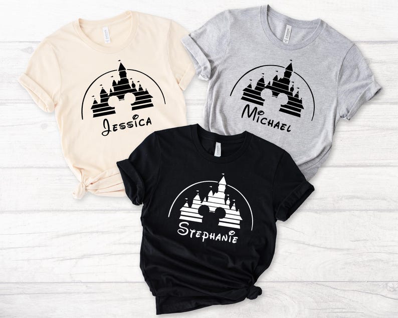 May include: Three t-shirts featuring a black and white graphic of Cinderella's castle with Mickey Mouse ears. The shirts are beige, grey, and black. The text on the shirts reads "Jessica", "Michael", and "Stephanie".