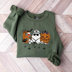 May include: Olive green sweatshirt featuring a Halloween-themed graphic. The design includes a ghost wearing a nurse's hat and sunglasses, a jack-o'-lantern, a spider, a bat, and medical equipment. The graphic is in shades of white, orange, black, and red.