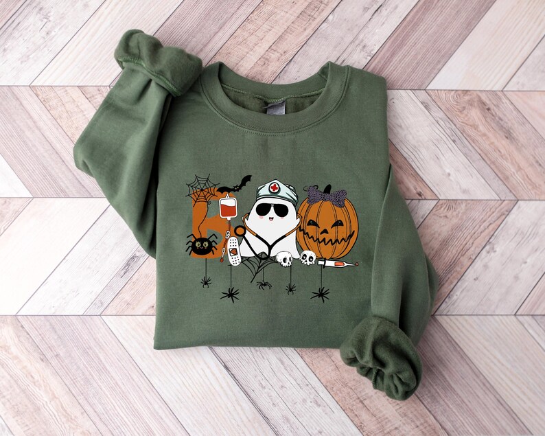 May include: Olive green sweatshirt featuring a Halloween-themed graphic. The design includes a ghost wearing a nurse's hat and sunglasses, a jack-o'-lantern, a spider, a bat, and medical equipment. The graphic is in shades of white, orange, black, and red.