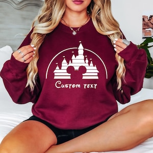 May include: Burgundy sweatshirt with a white graphic of Cinderella's castle and Mickey Mouse ears. The text "Custom Text" is below the graphic.