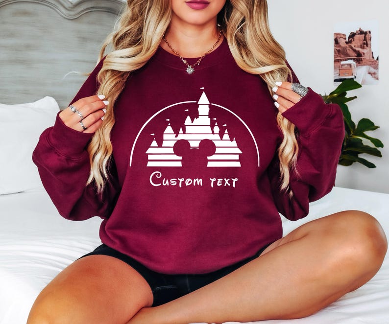 May include: Burgundy sweatshirt with a white graphic of Cinderella's castle and Mickey Mouse ears. The text "Custom Text" is below the graphic.