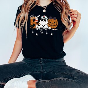 May include: Black t-shirt featuring a Halloween-themed design. The design includes the word "BOO" with a spider web, a ghost wearing a nurse's hat, and a carved pumpkin with a bow. The shirt is worn by a person.