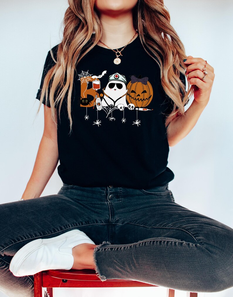 May include: Black t-shirt featuring a Halloween-themed design. The design includes the word "BOO" with a spider web, a ghost wearing a nurse's hat, and a carved pumpkin with a bow. The shirt is worn by a person.
