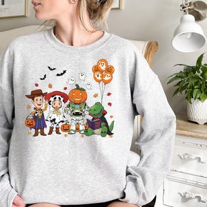 以下が含まれることがあります： ハロウィーンをテーマにしたグラフィックが特徴のライトグレーのスウェットシャツ。カウボーイ、ダルメシアン、カボチャ、恐竜など、仮装した漫画のキャラクターが登場します。オレンジ色の風船と落ち葉がデザインを完成させています。
