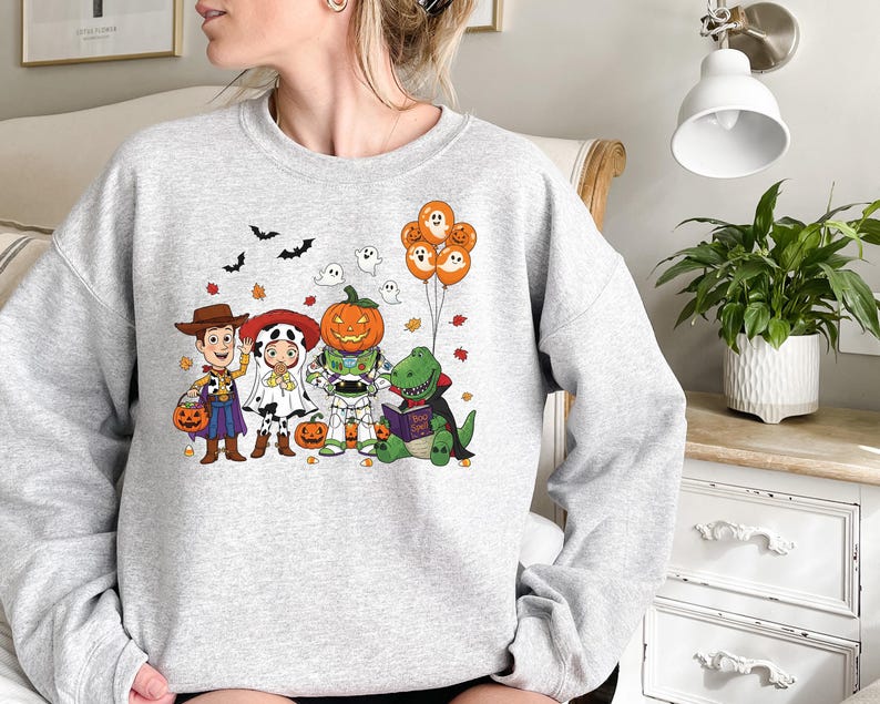 以下が含まれることがあります： ハロウィーンをテーマにしたグラフィックが特徴のライトグレーのスウェットシャツ。カウボーイ、ダルメシアン、カボチャ、恐竜など、仮装した漫画のキャラクターが登場します。オレンジ色の風船と落ち葉がデザインを完成させています。