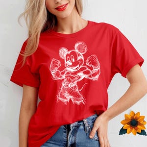 May include: A bright red short-sleeved t-shirt featuring a white line-art graphic of a muscular cartoon mouse character flexing its arms. The character is depicted in a sketch style with visible pencil-like strokes, wearing shorts and a belt.