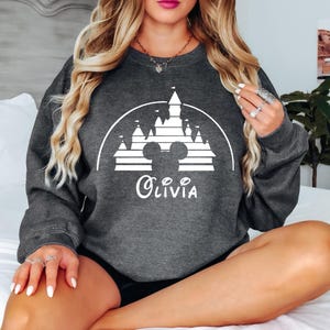 May include: A dark grey sweatshirt featuring a white silhouette of the Disney castle and Mickey Mouse ears. The text "Olivia" is printed below the image.
