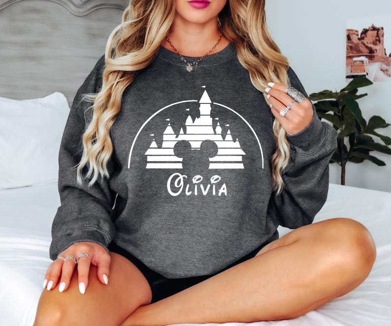 May include: A dark grey sweatshirt featuring a white silhouette of the Disney castle and Mickey Mouse ears. The text "Olivia" is printed below the image.