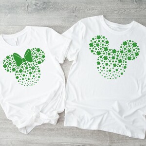 May include: Two white t-shirts with green shamrock designs. One shirt features a shamrock Mickey Mouse head, while the other has a shamrock Minnie Mouse head with a bow.