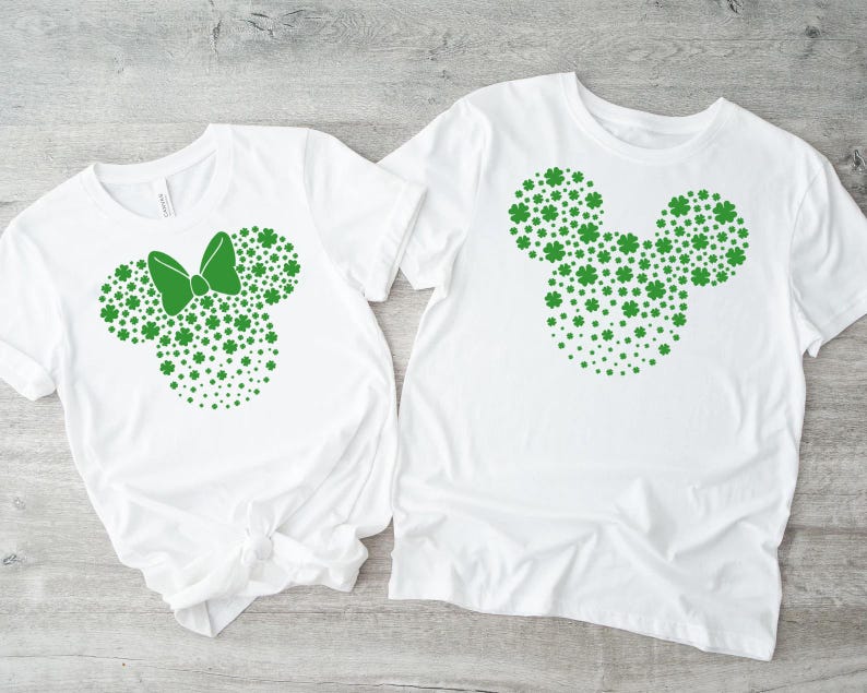 May include: Two white t-shirts with green shamrock designs. One shirt features a shamrock Mickey Mouse head, while the other has a shamrock Minnie Mouse head with a bow.