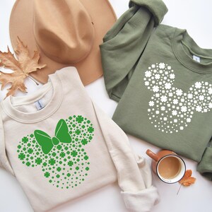 May include: Two cream colored sweatshirts with a green shamrock design in the shape of Mickey Mouse's head. One sweatshirt has a green bow on the design.