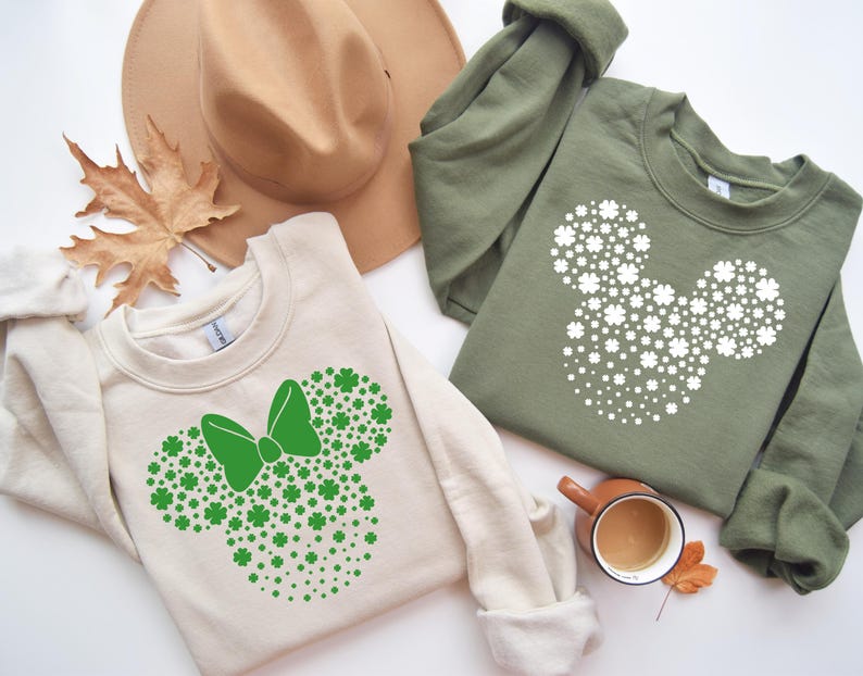 May include: Two cream colored sweatshirts with a green shamrock design in the shape of Mickey Mouse's head. One sweatshirt has a green bow on the design.