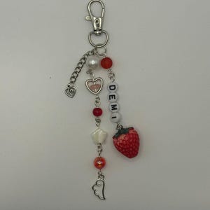 May include: A handmade keychain with a silver clasp and chain. The keychain features red and white beads, a heart charm, a star charm, a wing charm, and a red strawberry charm. The letters "DEMI" are spelled out with white letter beads.