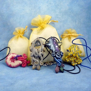 Puede incluir: Cuatro collares hechos a mano de arcilla polim&eacute;rica con dise&ntilde;os de tem&aacute;tica oce&aacute;nica. Los collares se muestran en bolsas de tul amarillo. Los dise&ntilde;os incluyen un pulpo gris con una esfera de reloj, un calamar morado y azul, un pulpo amarillo con anillos azules y un nautilus rosa y blanco.
