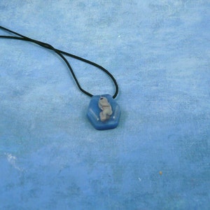 Puede incluir: Un colgante hexagonal azul con una pequeña figura blanca en su interior. El colgante está en un cordón negro.