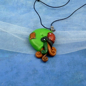 Puede incluir: Un colgante de arcilla polim&eacute;rica verde, marr&oacute;n y rojo con un dise&ntilde;o cosido y un collar de cord&oacute;n negro. El colgante tiene forma de espiral con un peque&ntilde;o ojo negro.