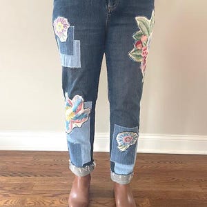 May include: Dark wash denim jeans with floral patches in shades of pink, blue, and green. The jeans have a relaxed fit and are cuffed at the ankles. The patches are sewn onto the front of the jeans.