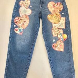 May include: Light blue denim jeans adorned with heart-shaped patches. The patches feature floral and patterned fabric in shades of pink, white, and yellow. The jeans have a classic five-pocket design and a straight leg cut.