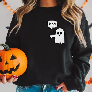 Puede incluir: Sudadera negra con un gráfico de fantasma blanco y la palabra "boo" en una burbuja de diálogo. Una calabaza naranja se sostiene en primer plano. Decoraciones temáticas de Halloween en el fondo.