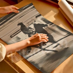 May include: A black and white print of a cowboy riding a horse, with motion blur. The print is being held by a person. A rolled-up poster is in the background. The print is on a wooden surface.