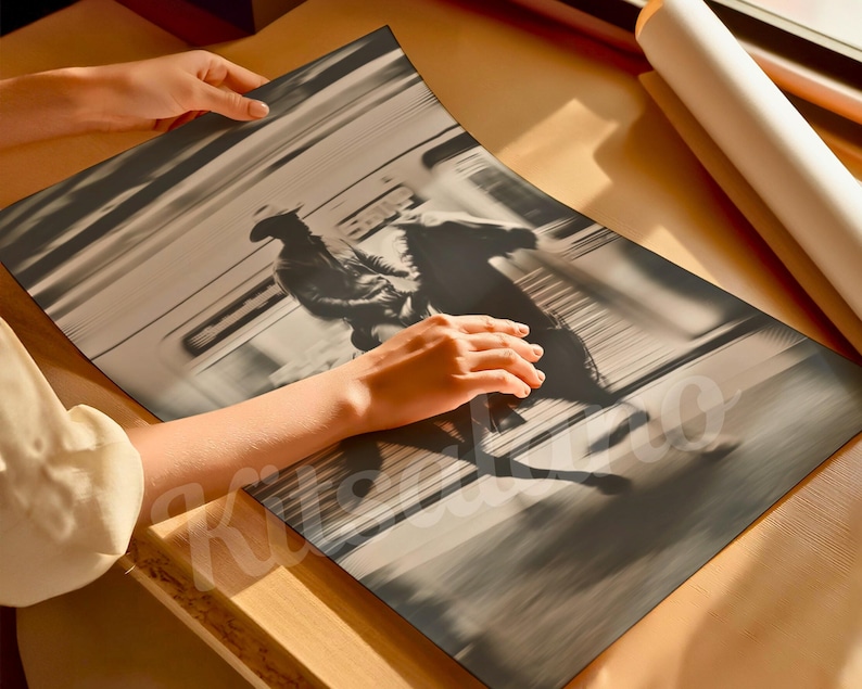 May include: A black and white print of a cowboy riding a horse, with motion blur. The print is being held by a person. A rolled-up poster is in the background. The print is on a wooden surface.