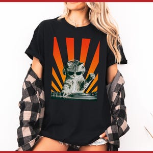 May include: Black t-shirt featuring a graphic of a cat wearing headphones and sunglasses, DJing on a turntable. The design includes orange and yellow sun rays in the background. The shirt is worn with a plaid shirt.