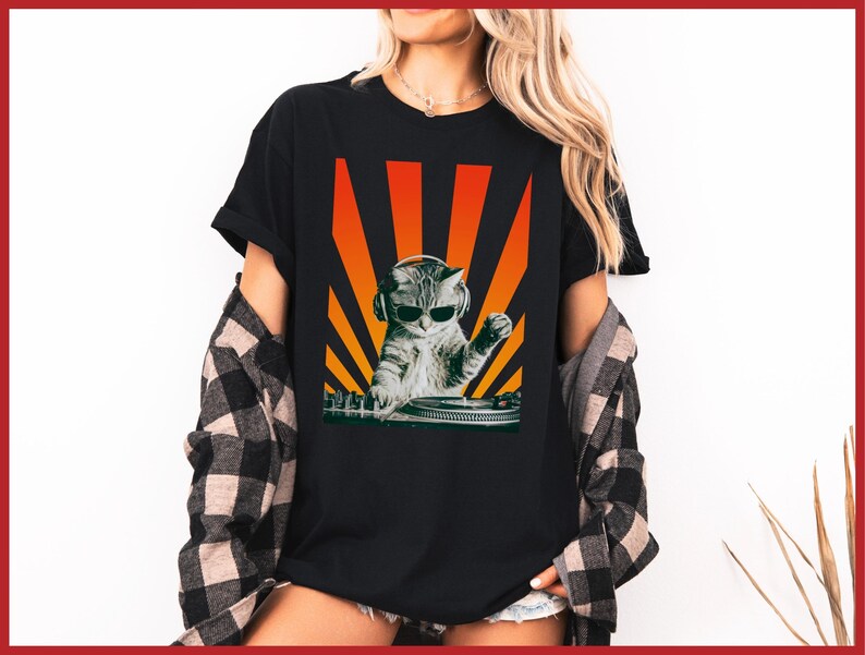 May include: Black t-shirt featuring a graphic of a cat wearing headphones and sunglasses, DJing on a turntable. The design includes orange and yellow sun rays in the background. The shirt is worn with a plaid shirt.