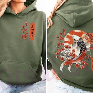 Pode incluir: Uma camisola com capuz verde-azeitona com um gr&aacute;fico frontal de flores de cerejeira vermelhas e texto em japon&ecirc;s. As costas apresentam uma grande ilustra&ccedil;&atilde;o de um peixe koi a nadar entre flores de cerejeira vermelhas e um sol, com texto em japon&ecirc;s.