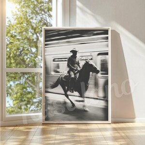 May include: Black and white framed print of a cowboy riding a horse in front of a blurred train. The cowboy wears a hat and is centered on the horse. The image is displayed in a room with a window and natural light.