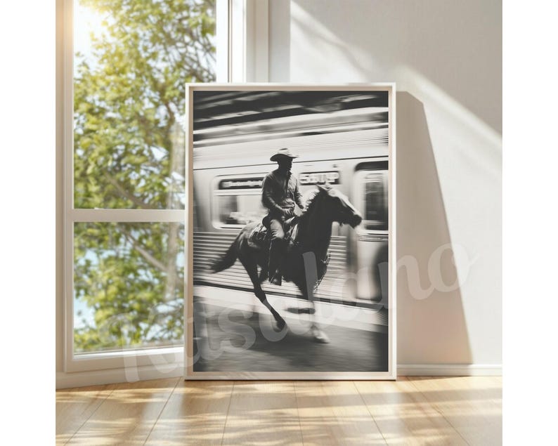 May include: Black and white framed print of a cowboy riding a horse in front of a blurred train. The cowboy wears a hat and is centered on the horse. The image is displayed in a room with a window and natural light.