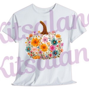 May include: White t-shirt featuring a floral pumpkin design. The pumpkin is white with a brown stem and is adorned with colorful flowers in shades of orange, pink, blue, and yellow. The design is centered on the front of the shirt.