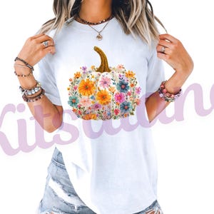 May include: White t-shirt featuring a floral pumpkin design. The pumpkin is white with a brown stem and is filled with colorful flowers in shades of orange, pink, blue, and yellow. The shirt is a casual style.