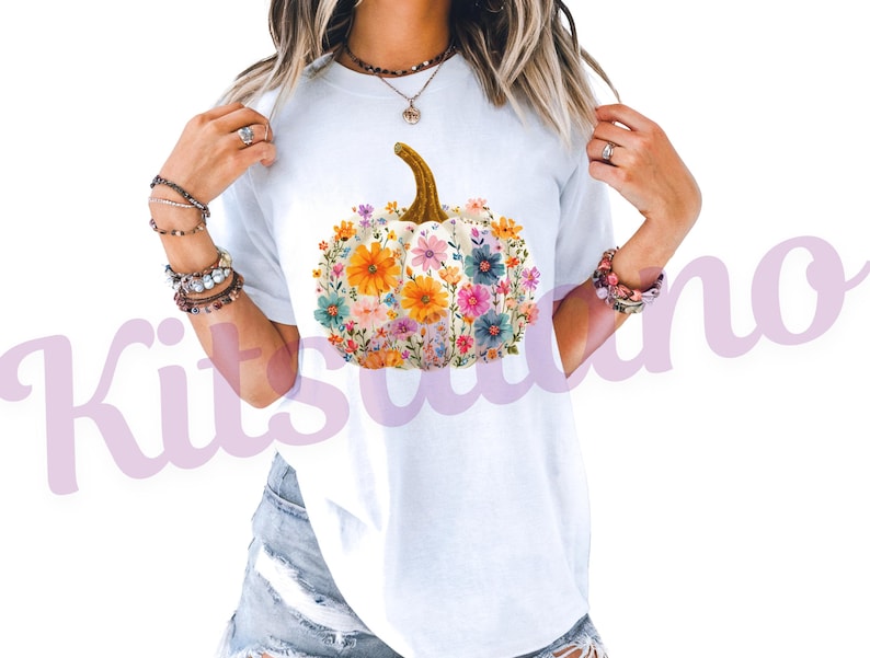 May include: White t-shirt featuring a floral pumpkin design. The pumpkin is white with a brown stem and is filled with colorful flowers in shades of orange, pink, blue, and yellow. The shirt is a casual style.