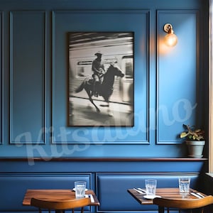 May include: A framed black and white photograph of a cowboy riding a horse, set against a blurred background of a train. The photograph hangs on a blue wall in a restaurant setting, with tables and chairs visible.
