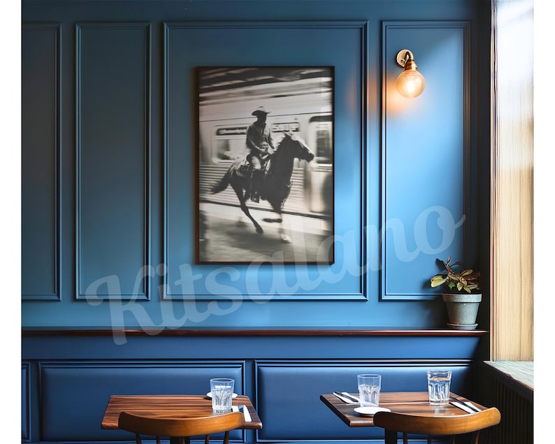 May include: A framed black and white photograph of a cowboy riding a horse, set against a blurred background of a train. The photograph hangs on a blue wall in a restaurant setting, with tables and chairs visible.