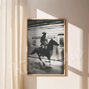 May include: Black and white framed print of a cowboy riding a horse past a train. The cowboy wears a hat and is seated on the horse. The train is blurred in the background, suggesting motion. The print is displayed on a white wall.