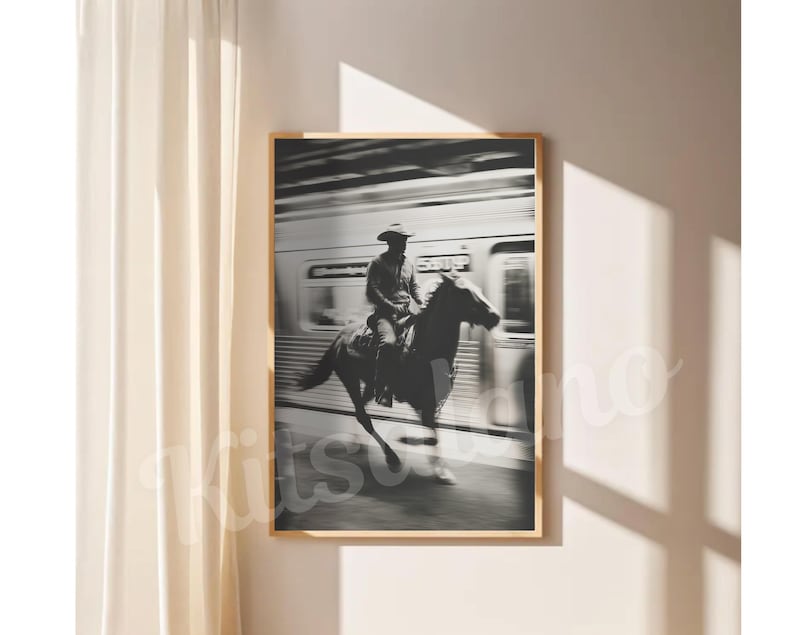 May include: Black and white framed print of a cowboy riding a horse past a train. The cowboy wears a hat and is seated on the horse. The train is blurred in the background, suggesting motion. The print is displayed on a white wall.
